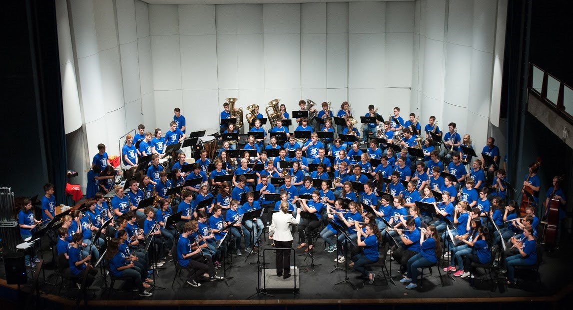 The Symphonic Band Camp Concert Band performs on stage in Durgin Concert Hall under the direction of Executive Director and Conductor Deb Huber featuring 150 high school band musicians in royal blue t-shirts with music instruments in hand.