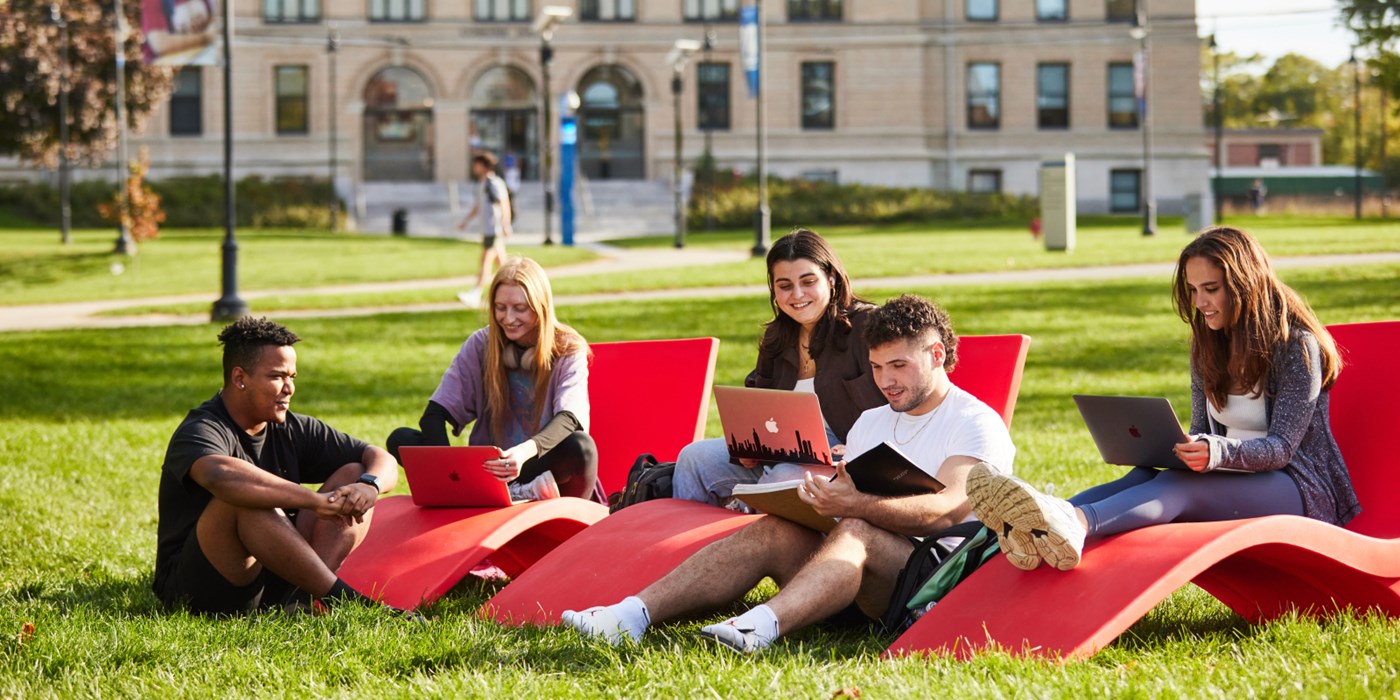 Five students sitting on lounge chairs and on the grass of south lawn with Coburn Hall in background