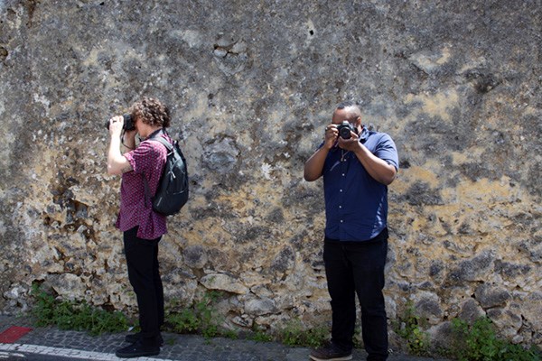 Two students each hold a camera and take a photo facing different directions against a large rock