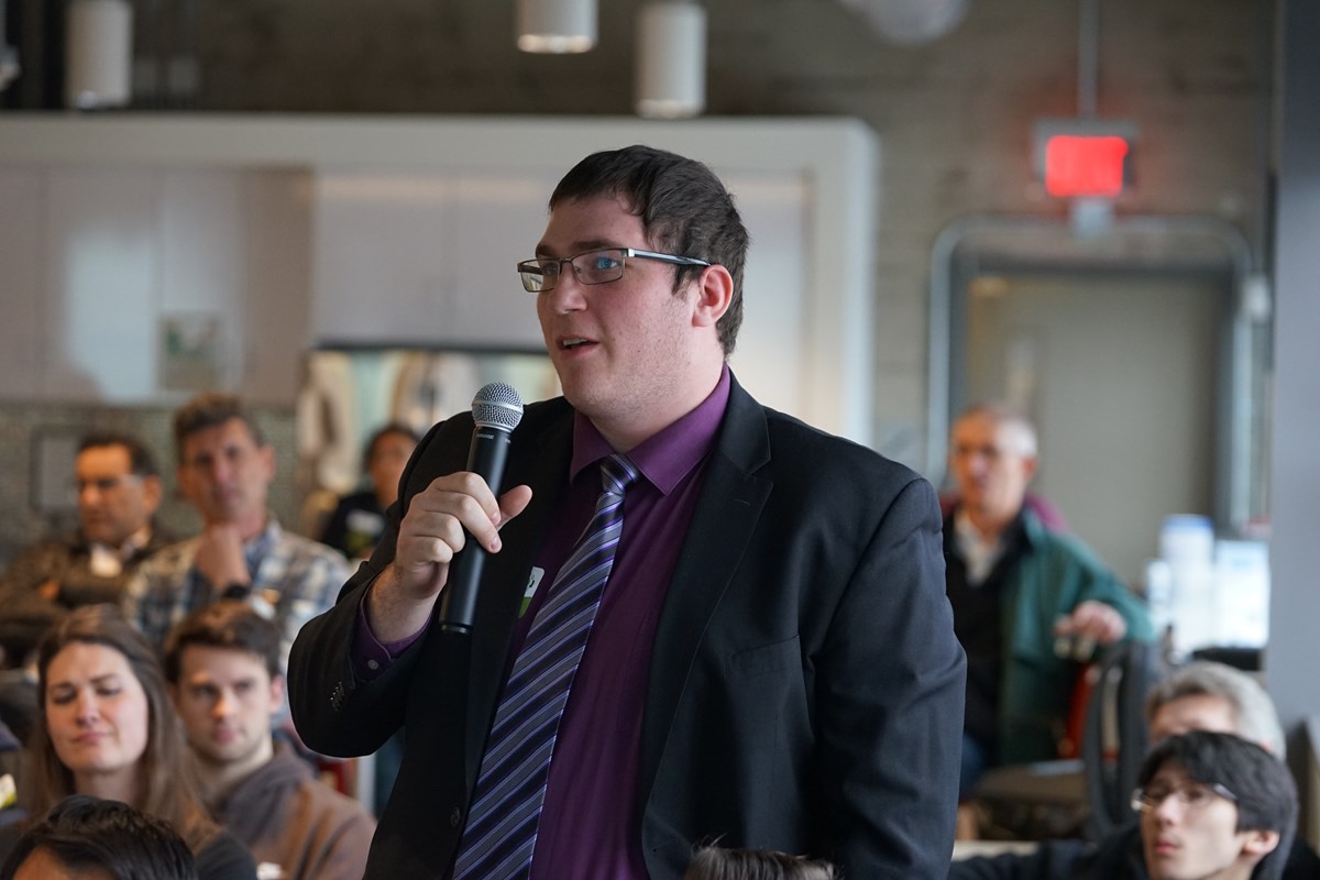 A person in a suit and tie and glasses holds a microphone and asks a question in an audience.