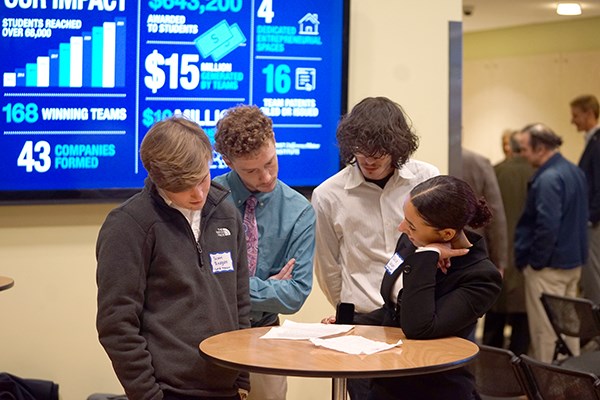 Three young men and a young woman look at papers on a table in front of a monitor
