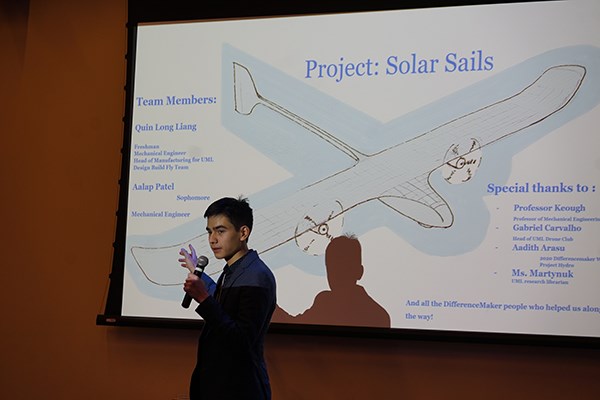 A student holding a microphone gestures to a screen with an airplane diagram behind him while making a presentation