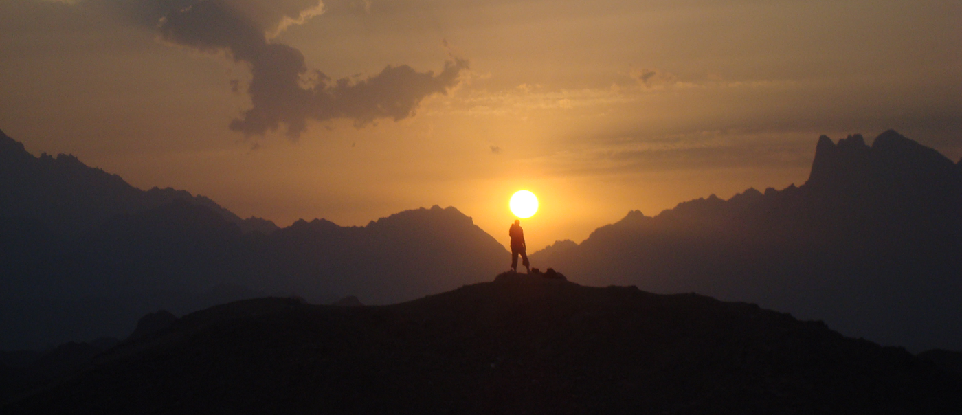 Person standing atop mountain with other mountains and sunset in distance.