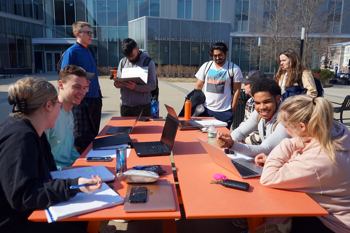 Nine students enjoy a nice day outside while working at an orange picnic table in front of a business school.