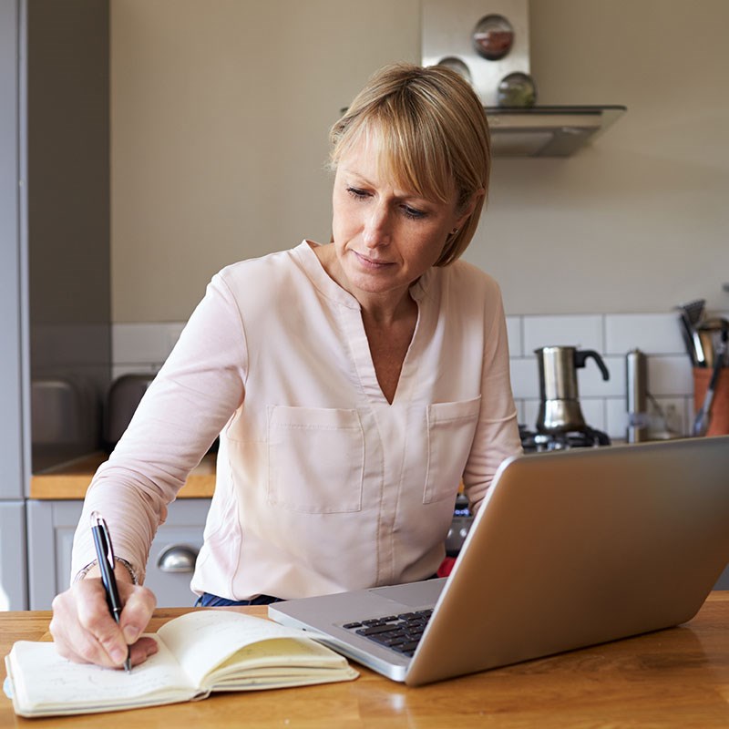 Woman seated at kitchen counter jots in notebook in front of laptop