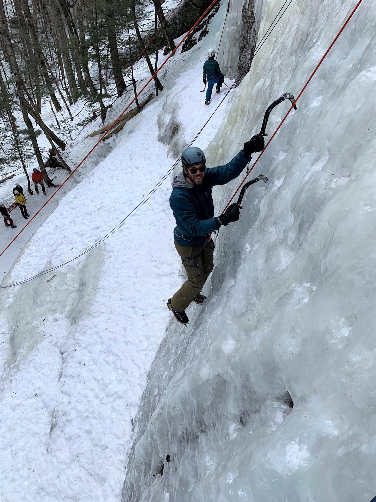 A person smiles while looking up and climbing ice with two ice tools in the ice.