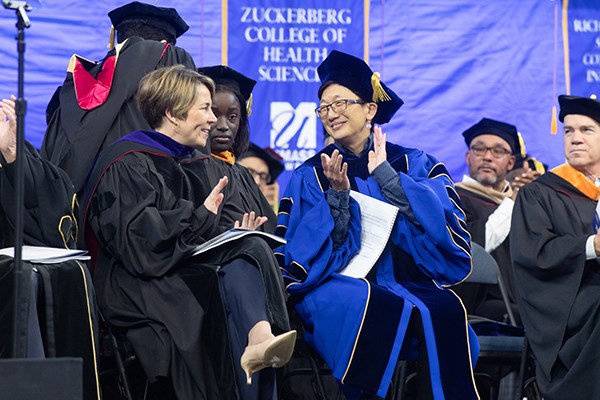 Two women clap while wearing academic regalia and sitting on a stage