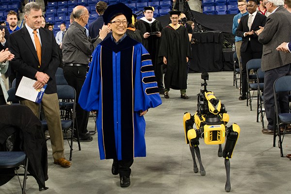 A woman in academic regalia walks down an aisle with a yellow robotic dog