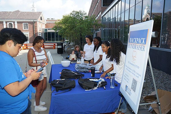 Four students in white T-shirts run a backpack fundraiser table outside with two customers