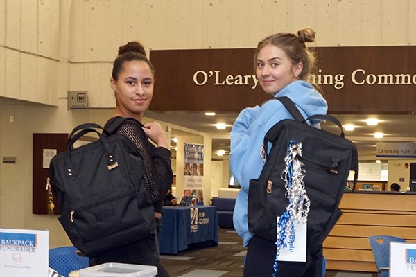 Two young women pose for a photo, each with a black backpack over their shoulder