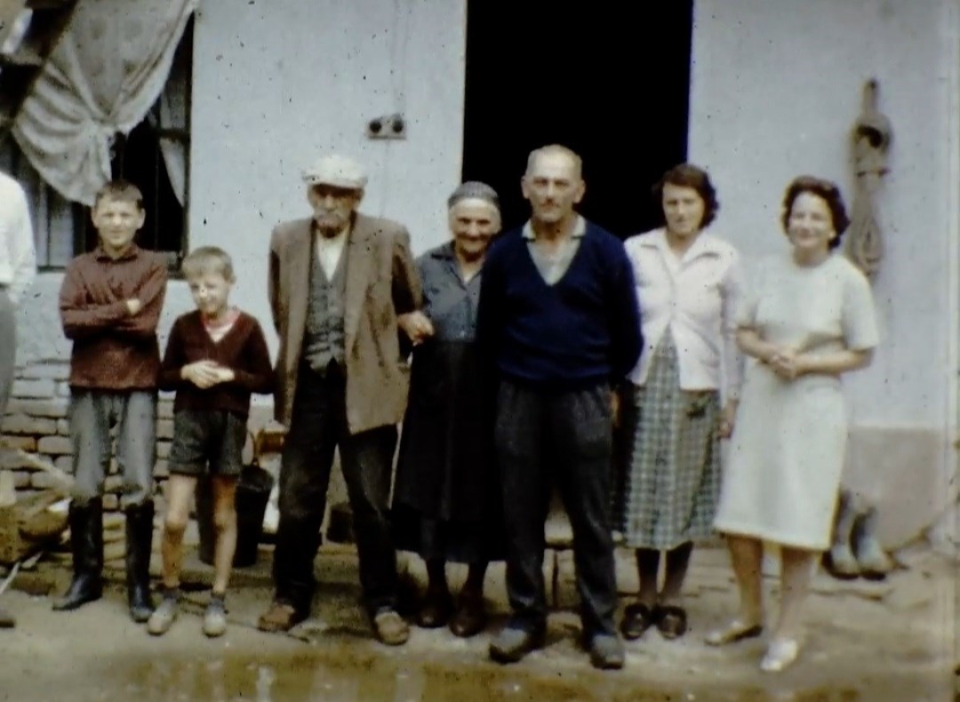 A group of people: five adults and two boys, standing together outside a house.