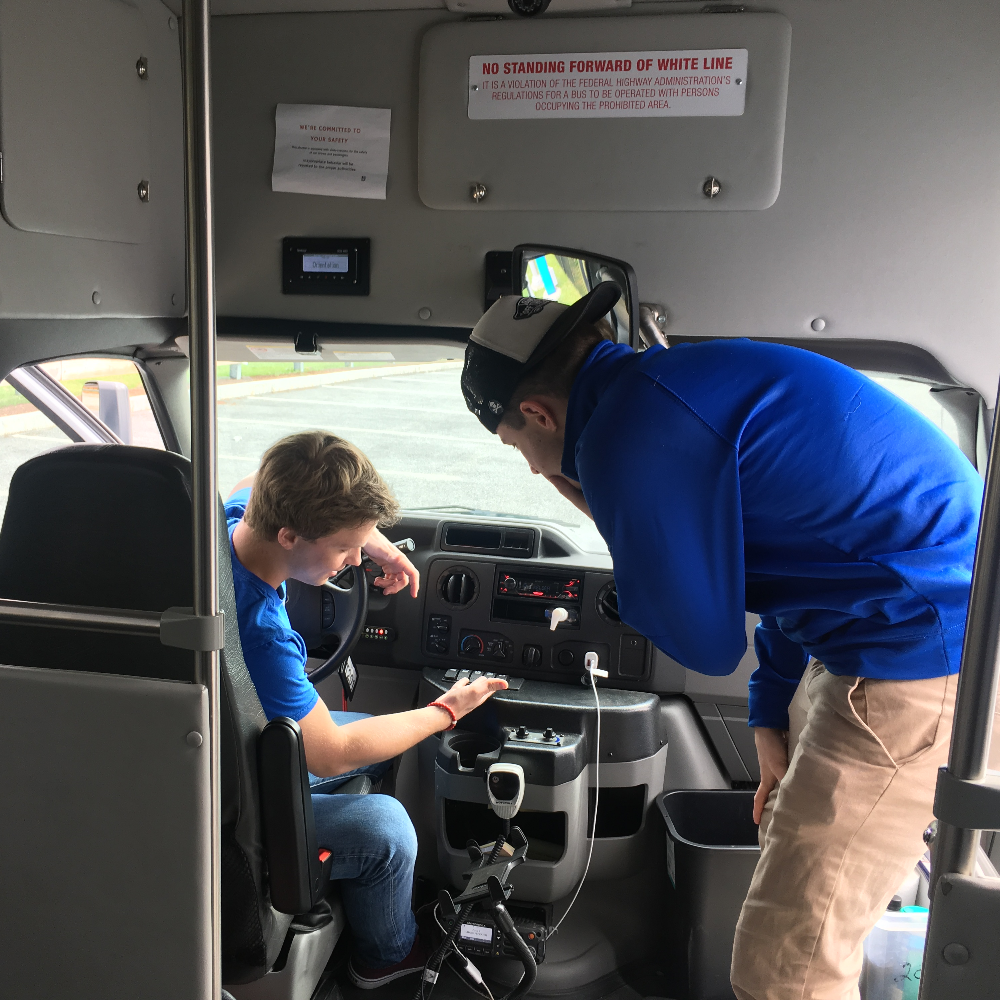 Two employees inside a shuttle bus, one is showing the other how to operate the switches. 