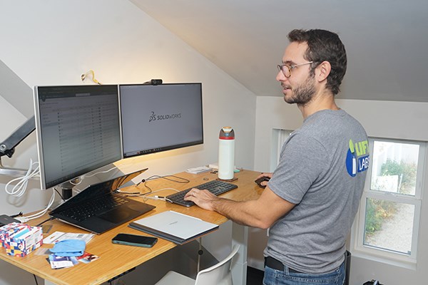 A man in glasses and a T-shirt works at a computer with two monitors
