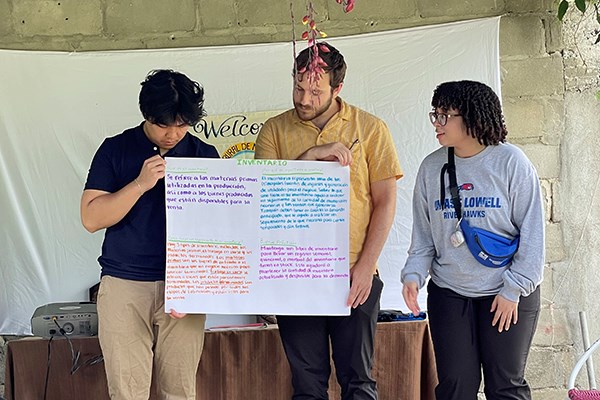 Two young men hold a poster board while a young woman makes a presentation outdoors