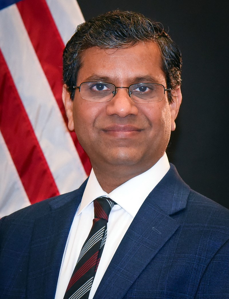 Ravi Mosurkal wearing a suit and glasses and smiling with an American flag behind him.