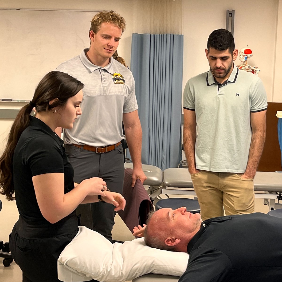 Three students stand around a patient lying on a table in the UMass Lowell physical therapy clinic.