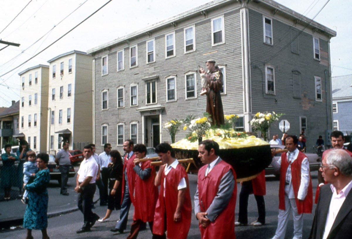 Procession at the Feast of St. Anthony: a group of people, many wearing red garments over their clothes, walk down a Lowell street in 1980 carrying a display with a statue of St. Anthony holding a child  with cars and buildings in the background.