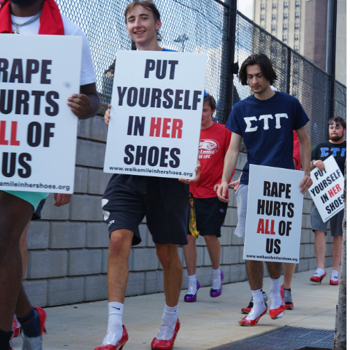 Young men walking across bridge wearing red heels holding signs that say "Put Yourself in Her Shoes"