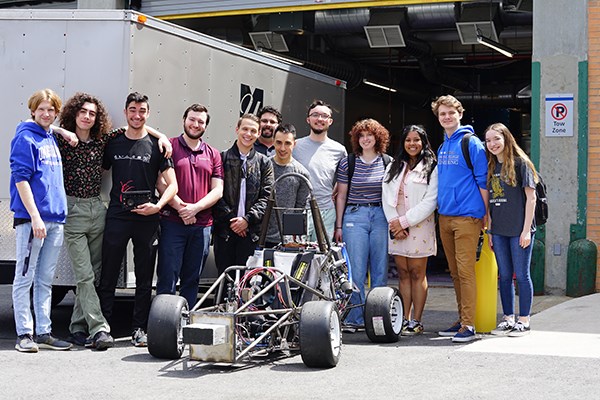 A dozen students pose for a photo with their formula-style race car they are building