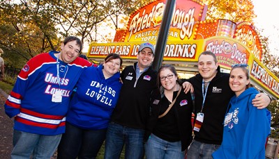Group of people in UMass Lowell clothes posing in front of a fried dough and carnical food stand.
