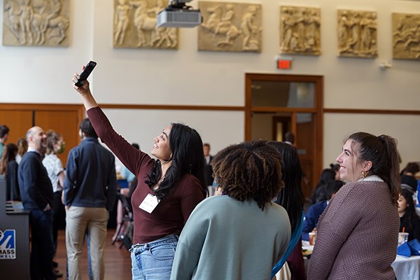 A young woman holds up her phone to take a selfie with three other students inside a function room