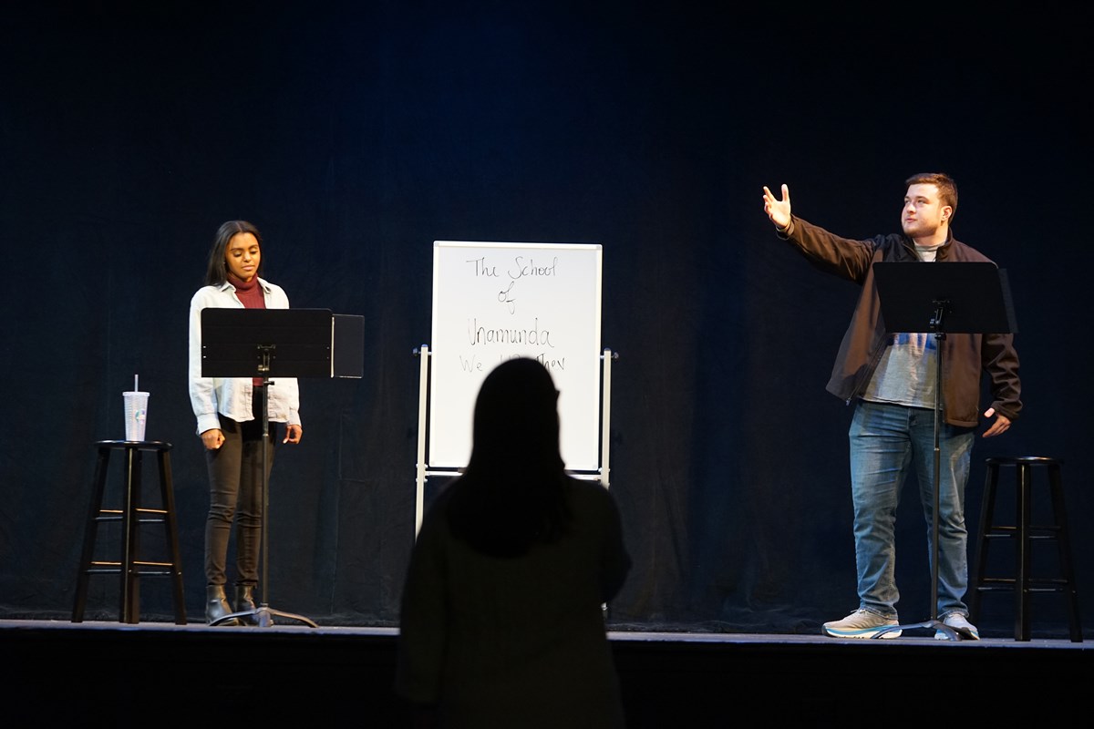 A young man gestures with his arm on stage next to a young woman. A silhoutte of head is between them.