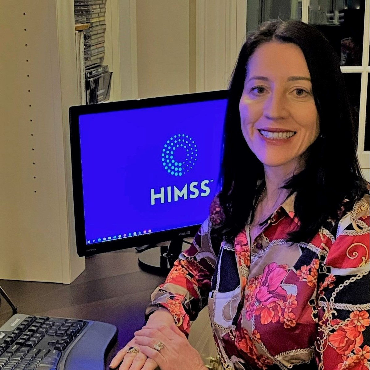 Toni Laracuente sits at a desk with a computer screen that displays the logo and letters HIMMS (Healthcare Information and Management Systems Society).