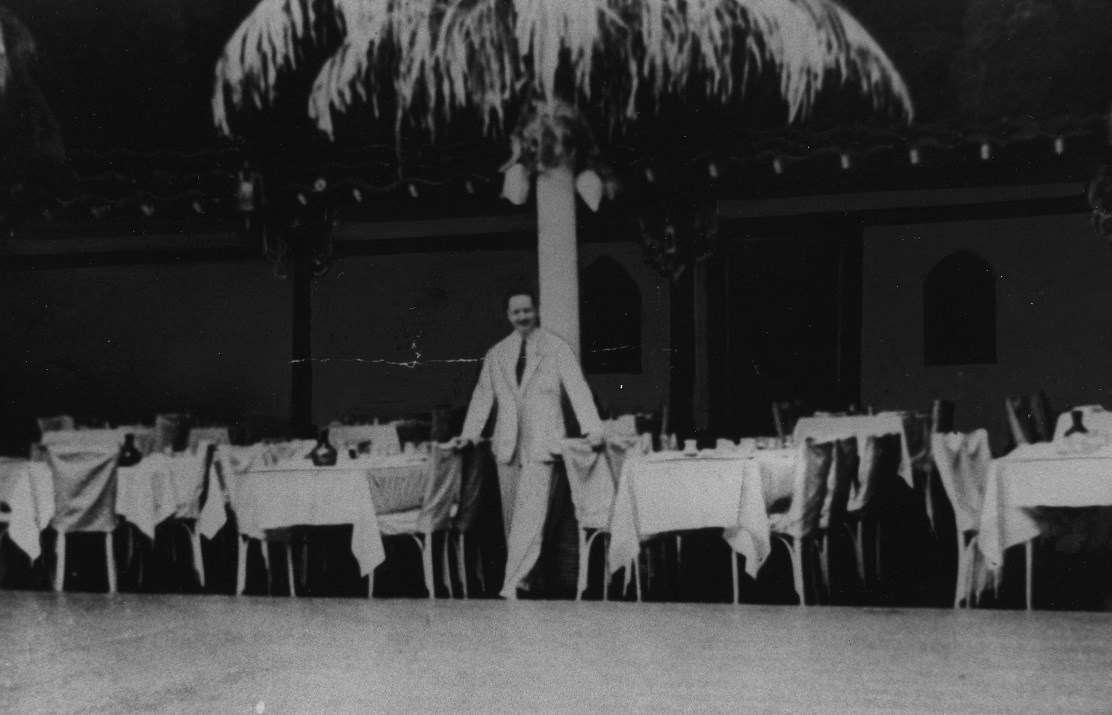 Black and white photo of person in white suit standing in front of palm tree with dining tables and chairs to each side.