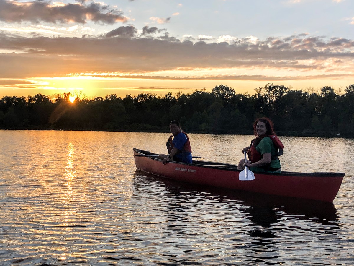 Two people smile in a canoe as the sun sets beyond