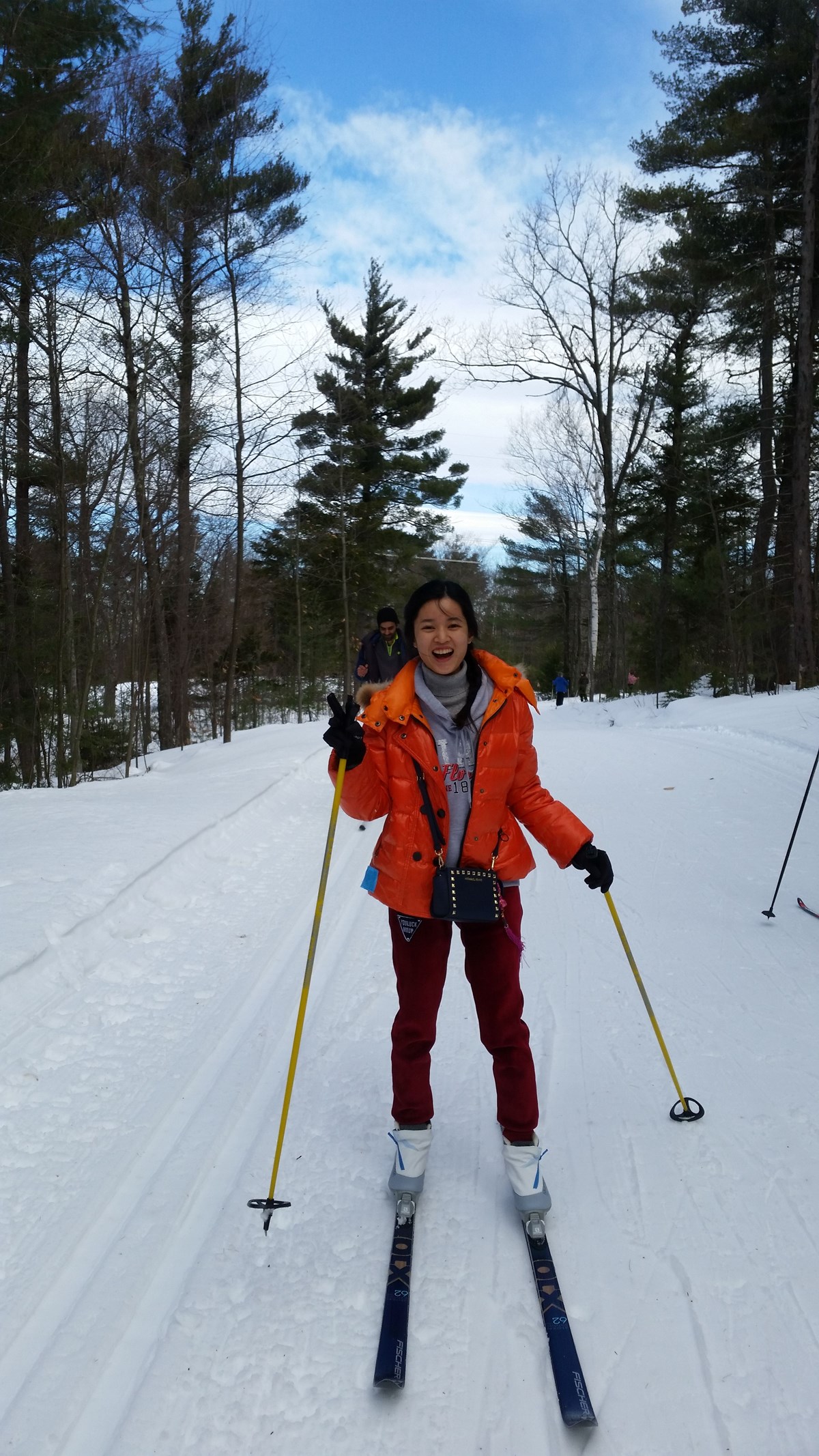a woman wearing an orange coat grins while doing cross country skiing on a groomed trail outside