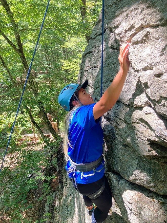 A woman wearing a helmet and harness climbs a rock and reaches up for a hold.