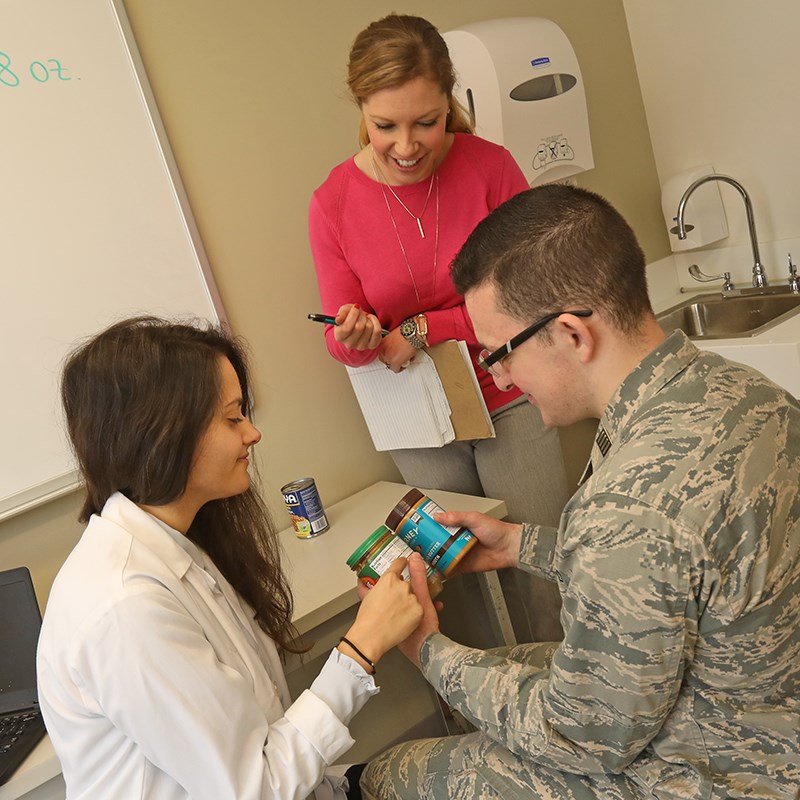 Two female dietetics students help a ROTC student compare labels on jars of nut butter.