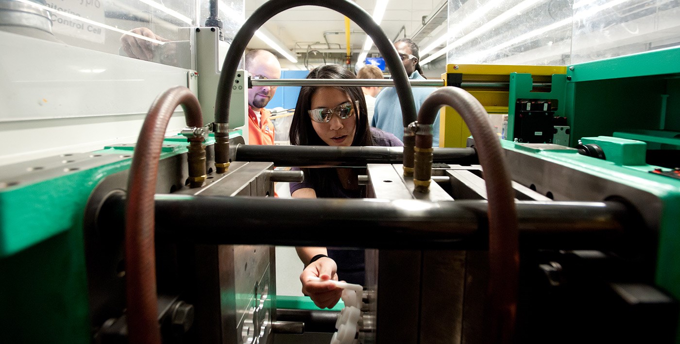A female UMass Lowell student seen through machinery in a Plastics Engineering lab.