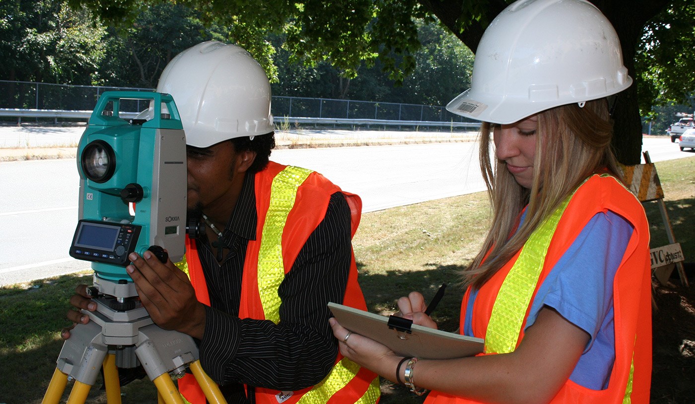 A male and female UMass Lowell student both wearing hard hats and surveying.