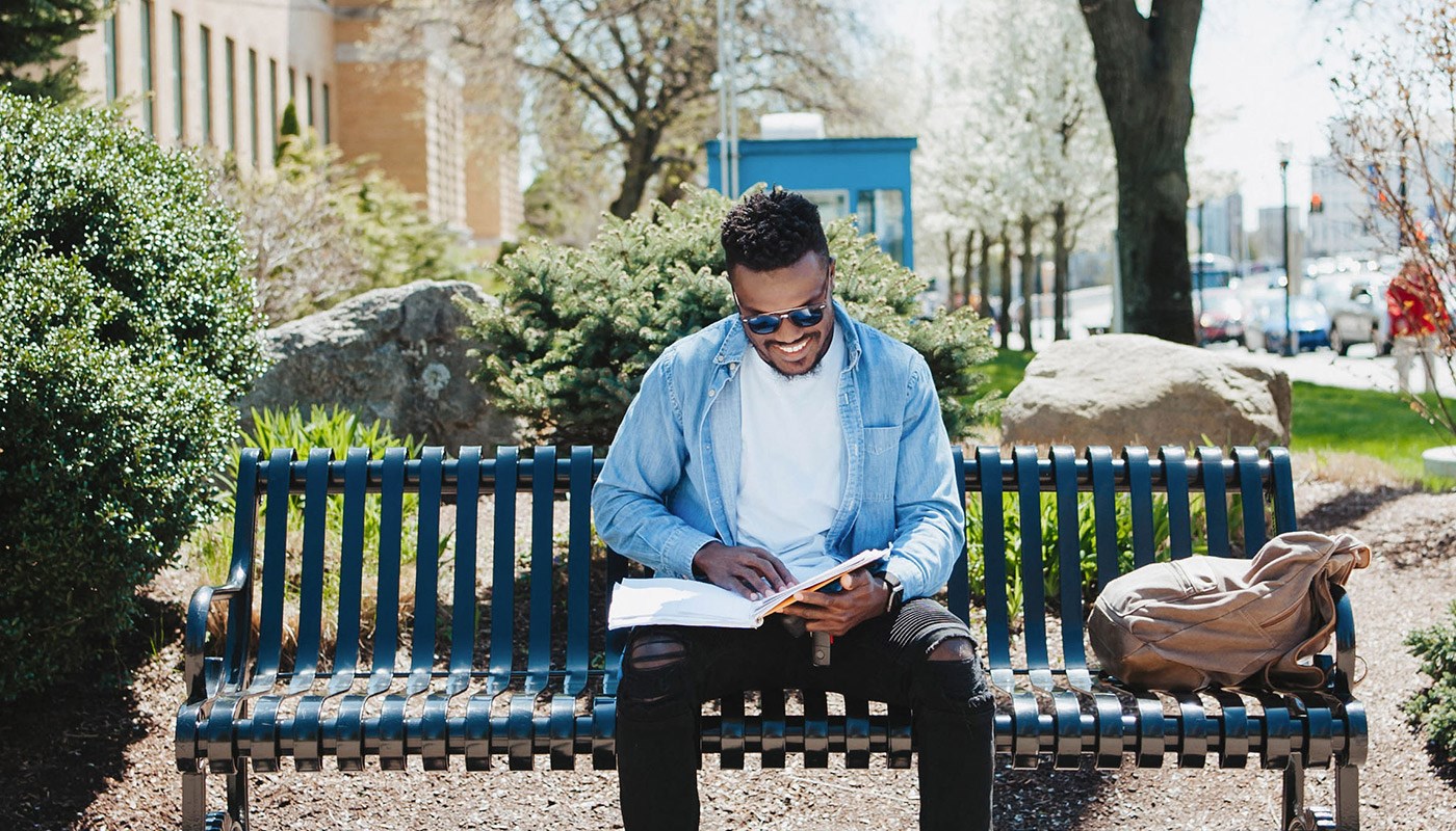Male student sitting on a bench reading a textbook on UMass Lowell's north campus