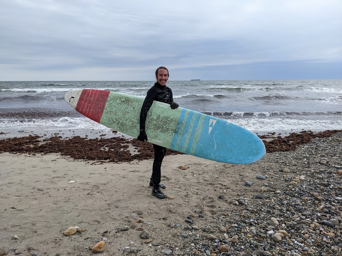A surfer stands on a beach holding a board smiling with the ocean and small waves behind.