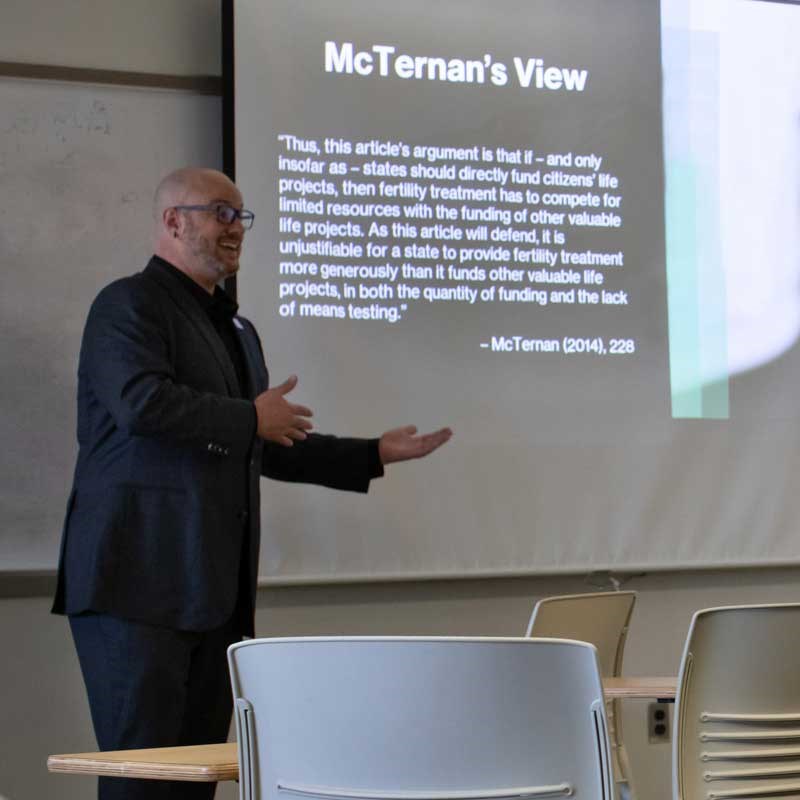 UMass Lowell professor Blake Hereth speaks before a classroom with a slide showing the words: McTernan's View