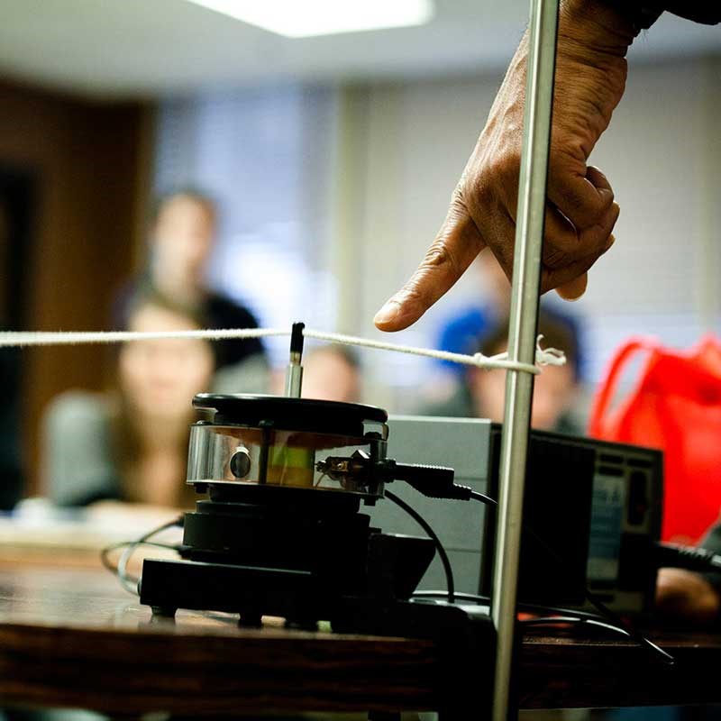 A finger points to a string vibrating in a UMass Lowell physics classroom