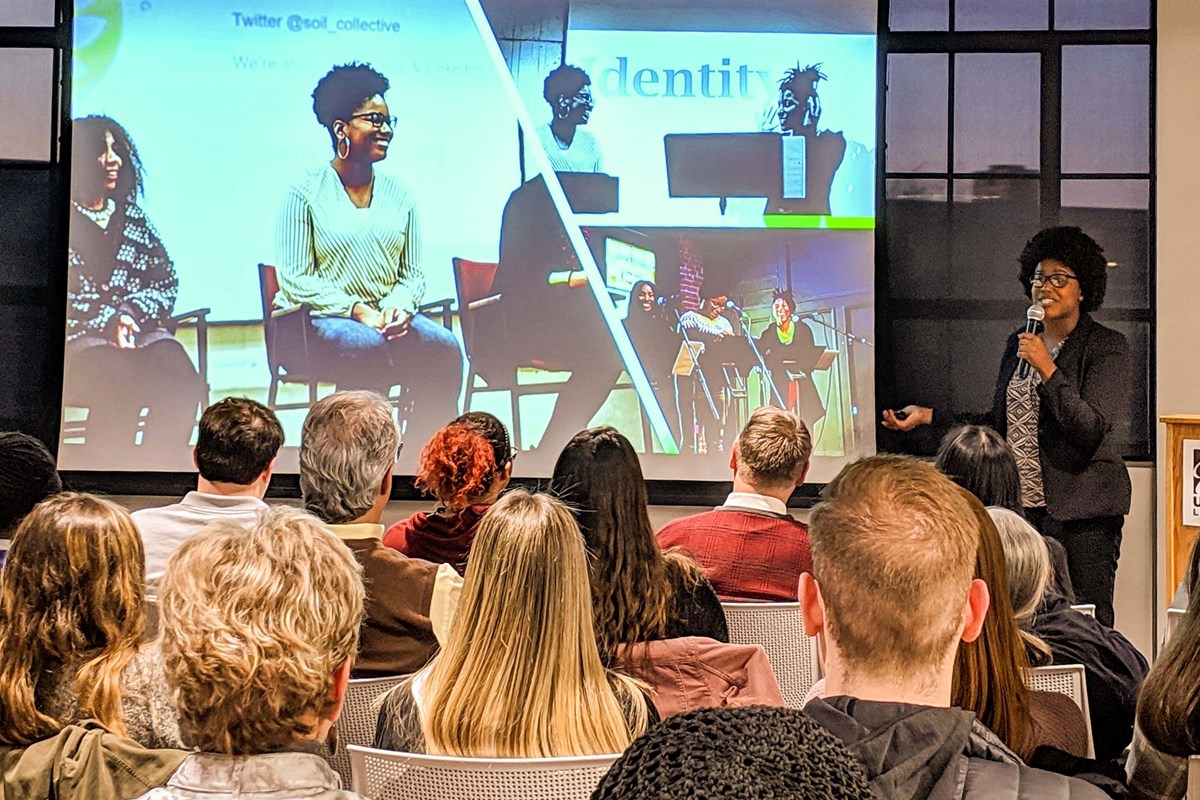 Person talking into a microphone in front of a seated audience with a screen featuring other people to the left.