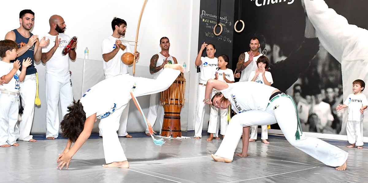 Two people demonstrating Capoeira—a dynamic combat game that melds play, fight, dance, acrobatics, music and ritual as others watch around them and another plays a drum.