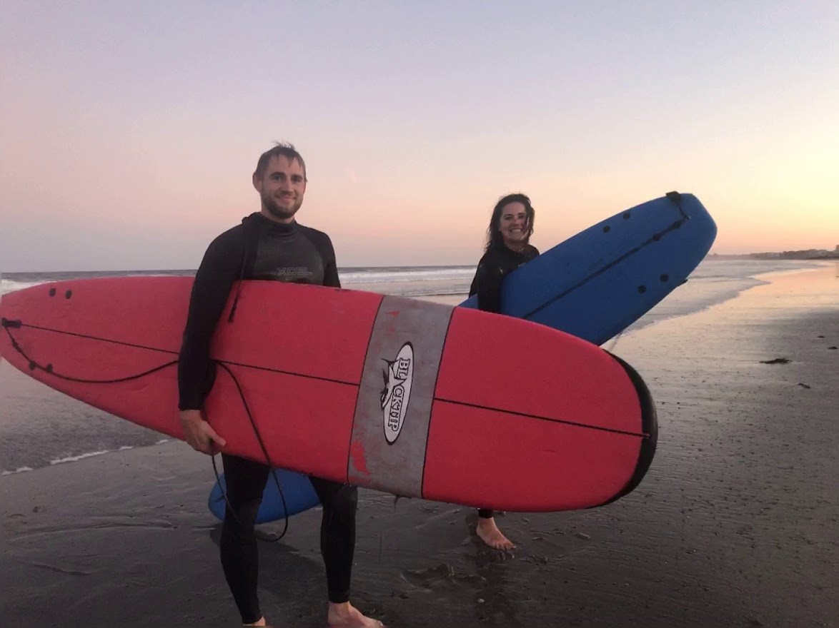 2 people in wetsuits stand on the beach with surfboards in hand.