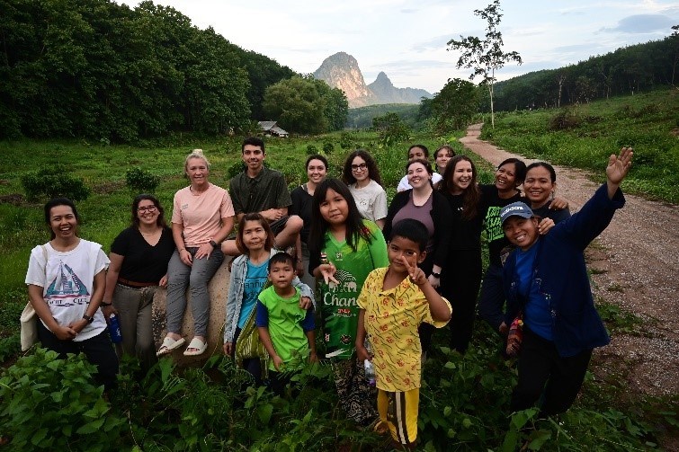 Group of students and local people smile and wave along dirt road with mountain in background