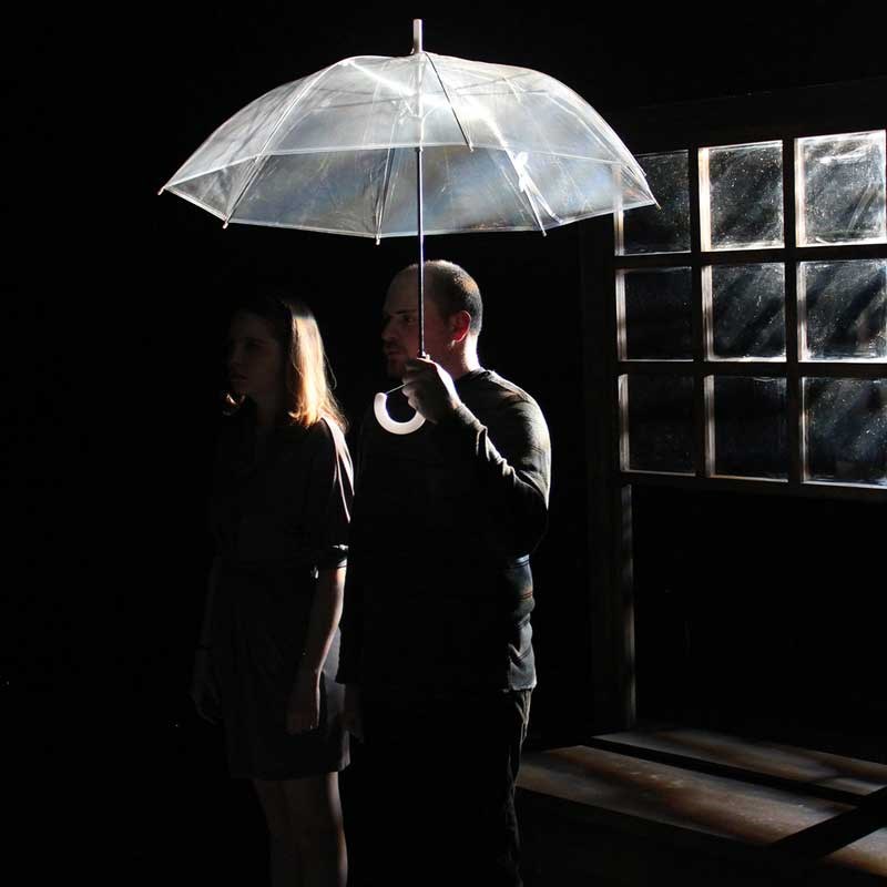 A male student holding an umbrella on stage next a female student with an window behind them in the dark during a UMass Lowell Theatre Arts production.
