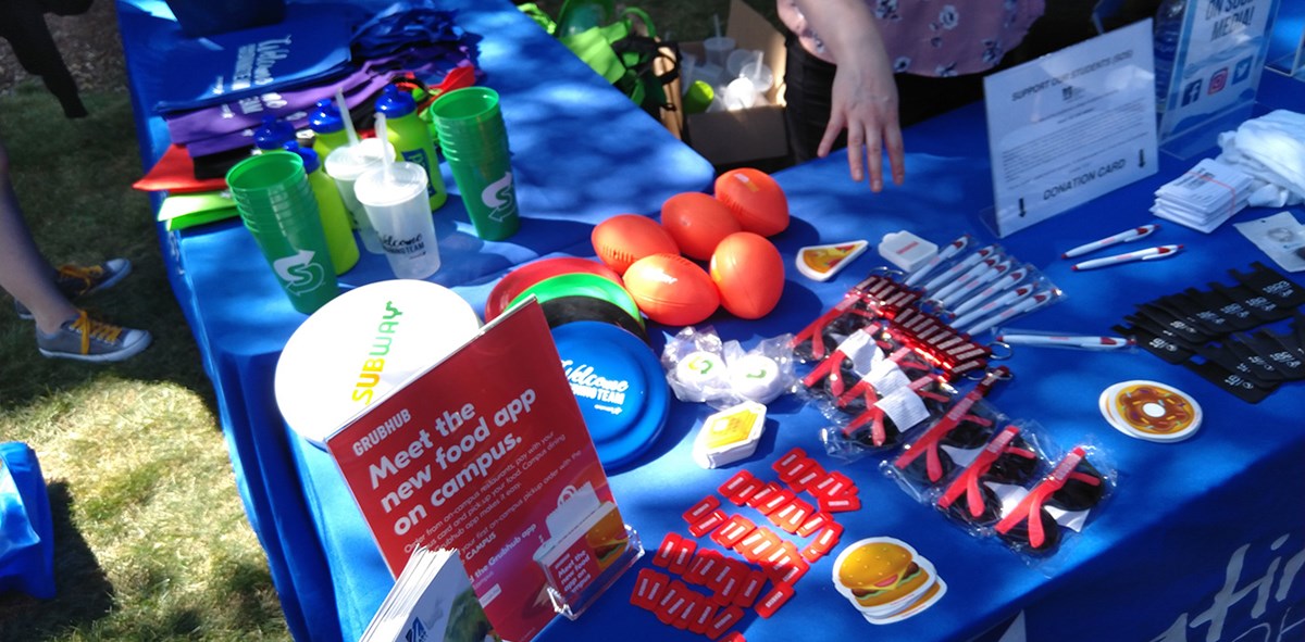 A table displaying UMass Lowell and other vendor branded swag including: frisbees, cups, sunglasses, pens, bags, balls and more.