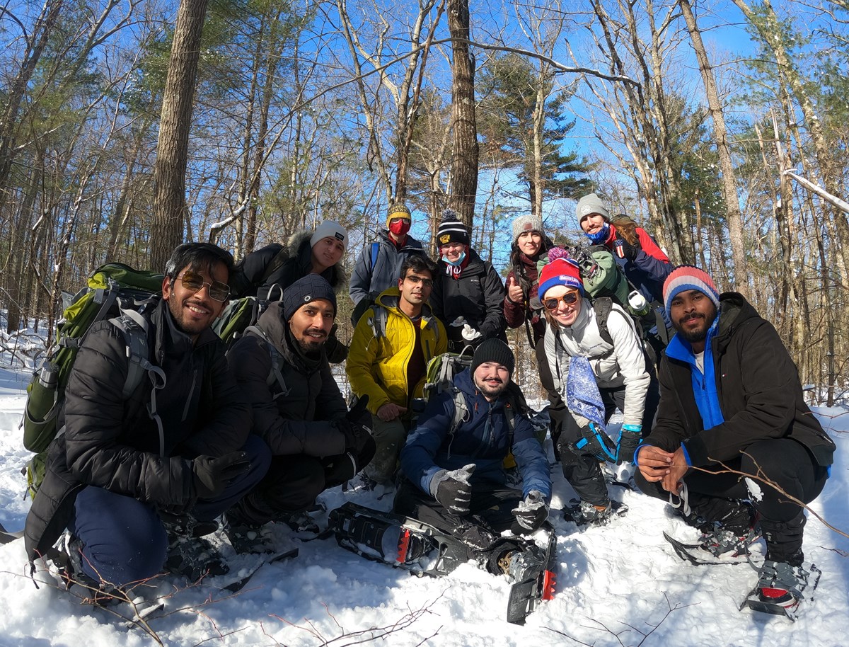A smiling group of people wearing snow-shoes and winter clothes in the snow with trees and a blue sky behind them.