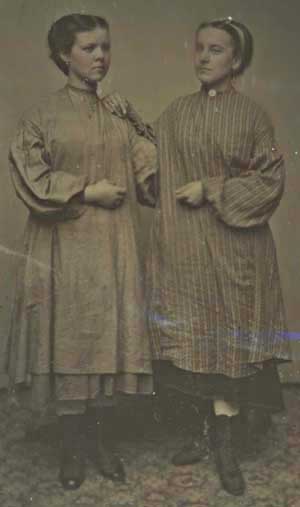 Dark photograph of 2 young women wear work smocks over street clothes. Showing there high top shoes.