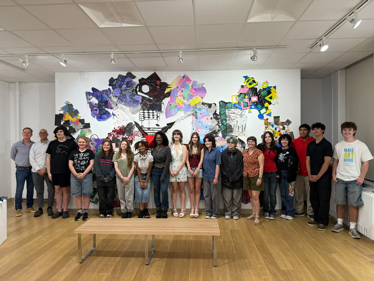 Faculty and students stand in a row for group picture in art gallery along a color wall of artwork.