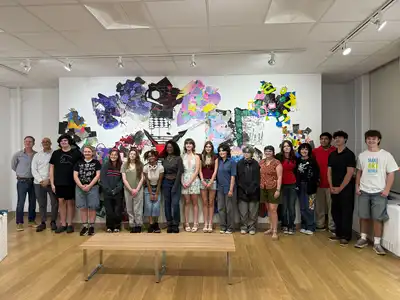 Faculty and students stand in a row for group picture in art gallery along a color wall of artwork.