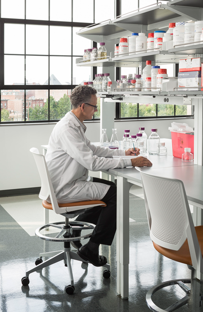 Man in white lab coat sitting at a desk with jars and other lab equipment on it as well as on shelves above him.