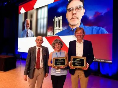 Three people stand on stage with two people on a screen behind them.